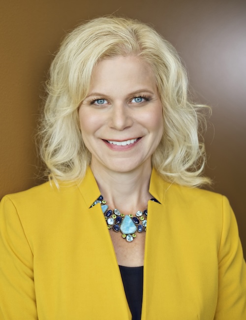 Kim Sponem – blonde hair, blue eyes, wearing yellow jacket and necklace
